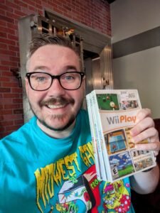 Selfie of man with beard and glasses holding video games, smiling at camera